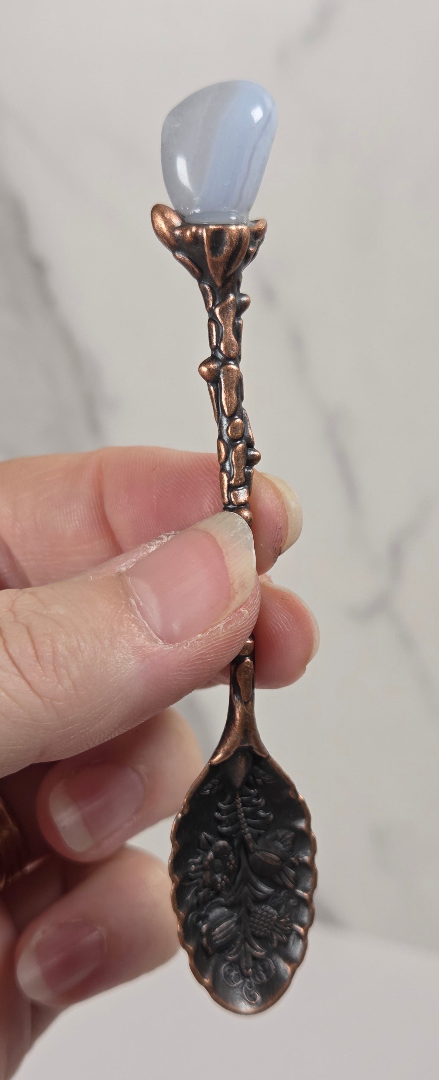 A hand holding a decorative spoon with a copper-toned leaf-shaped handle, a pale blue stone at the top, and a dark leaf-shaped bowl, set against a light marble-patterned background.