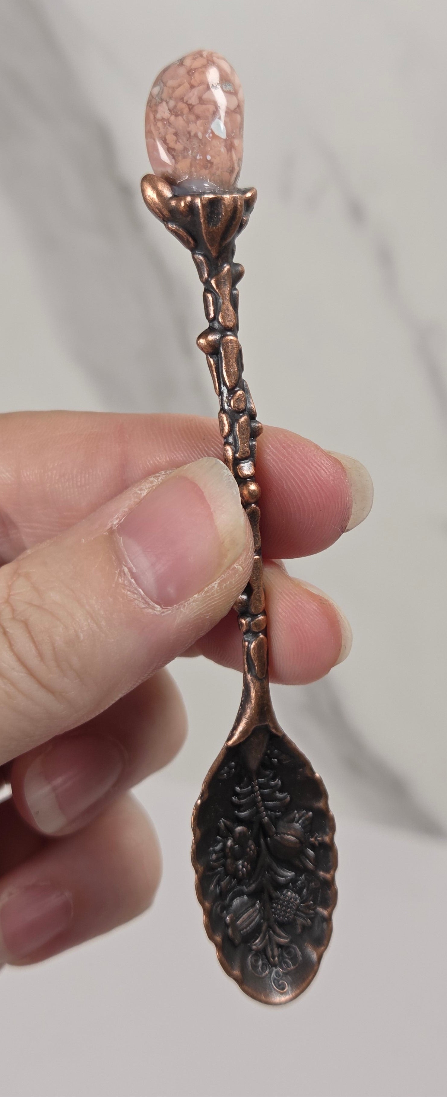 A hand holding a decorative copper spoon with a leaf-shaped bowl and a pink crystal gemstone at the top, set against a light gray marble background.