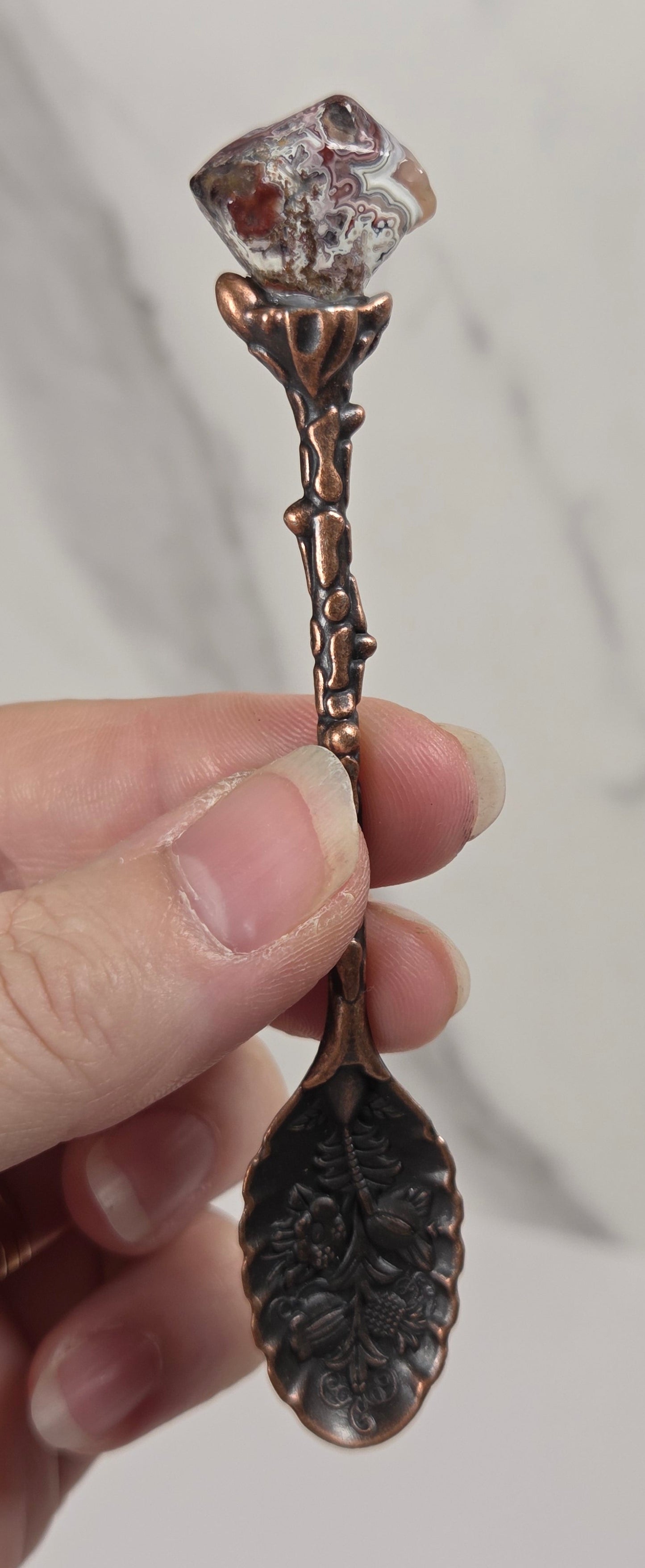 A hand holding a decorative copper spoon with a leaf-shaped bowl and a crystal gemstone at the top, set against a light gray marble background.