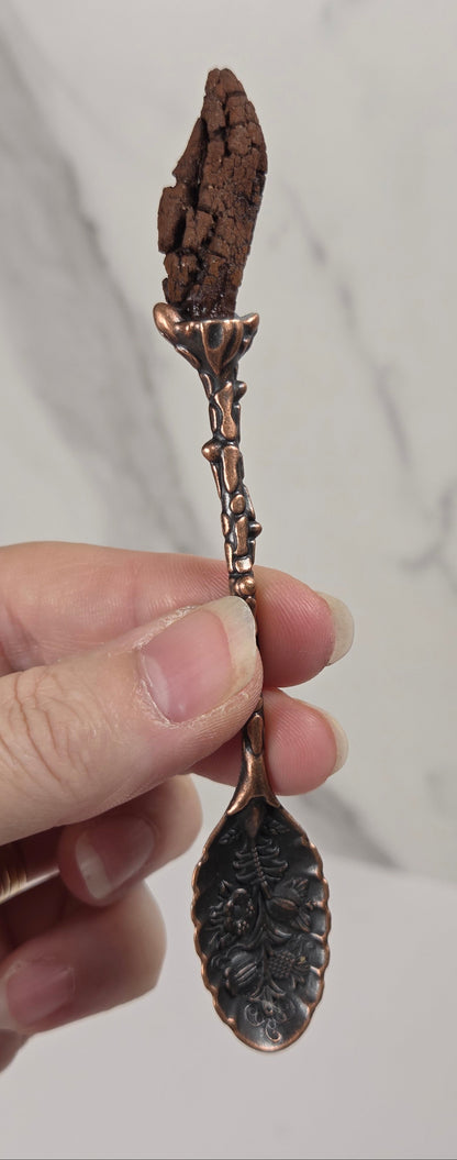 A hand holding a decorative copper-toned spoon with a textured, bark-like top and an embossed leaf-shaped handle, set against a light gray marble-patterned background.
