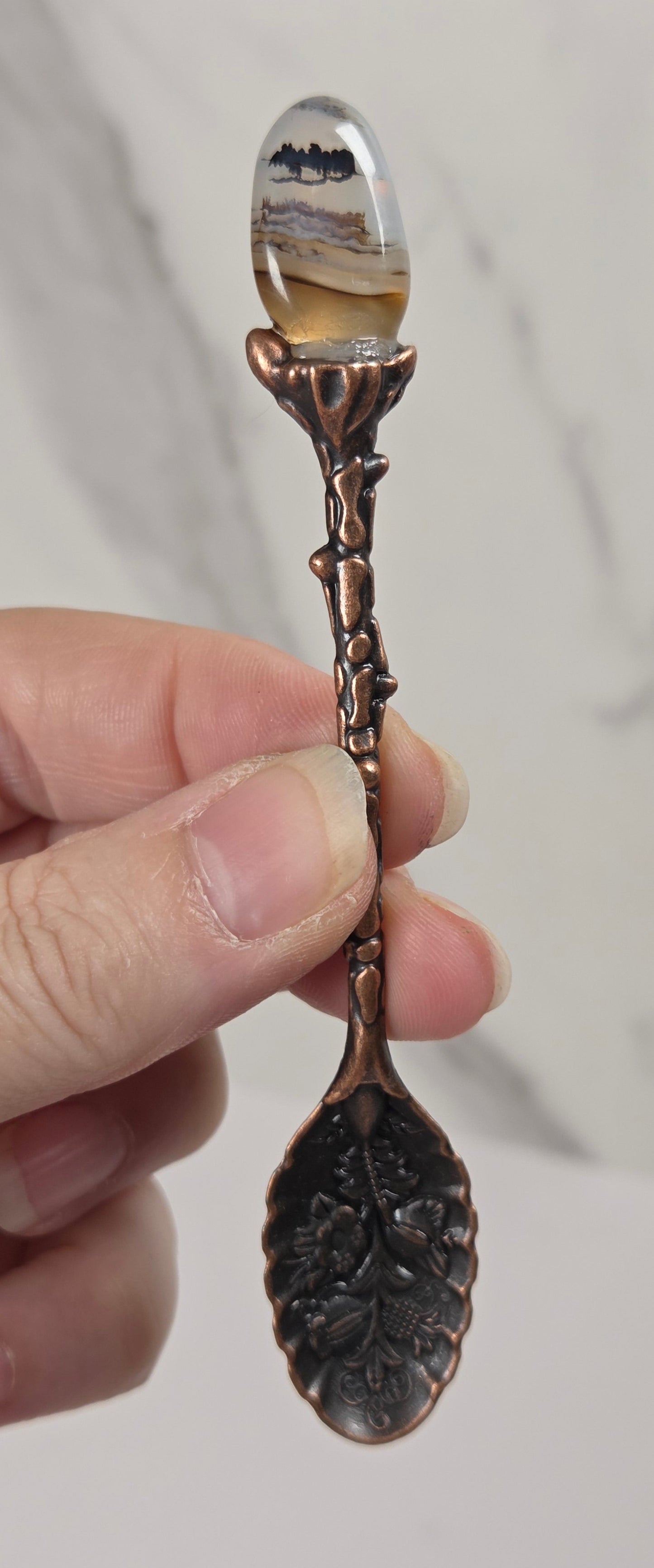 A hand holding a decorative spoon with a copper-colored twig handle, a leaf-shaped spoon bowl, and a clear oval top containing a landscape scene.