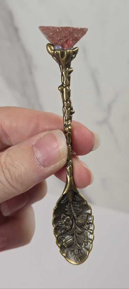 A hand holding a decorative antique-style brass spoon with a pink crystal point at the handle end, set against a light gray marble-patterned background.