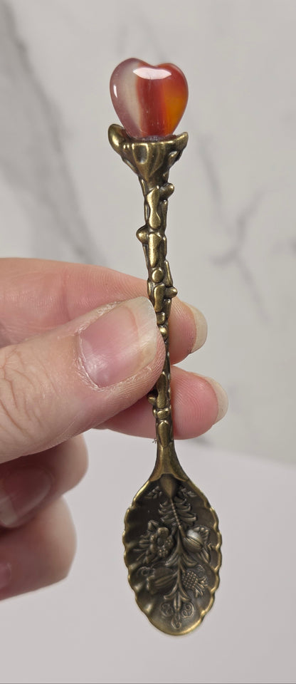 A hand holding a decorative antique-style metal spoon with a carved leaf-shaped bowl and a heart-shaped amber cabochon on the handle, set against a light gray marble-patterned background.