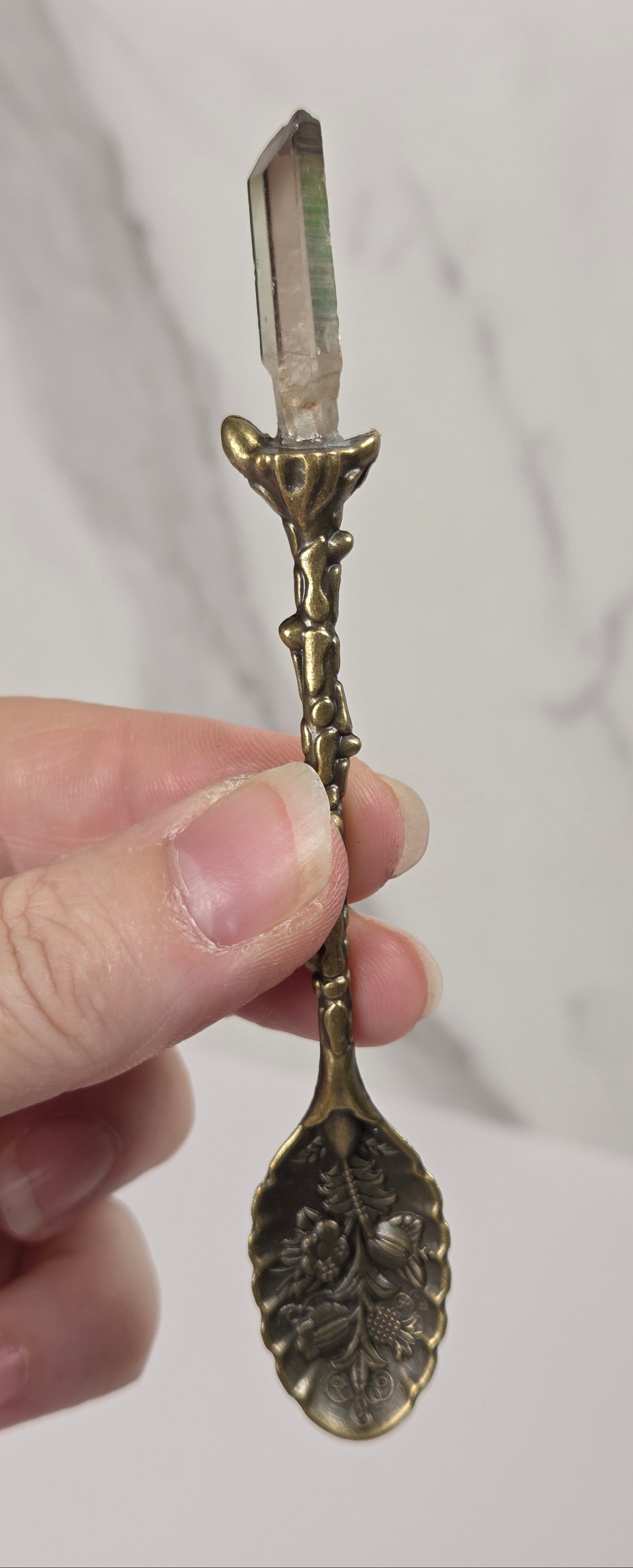 A hand holding a miniature ornate brass spoon turned into a crystal pointer wand with a pointed iridescent crystal at the top, set against a light gray marble background.