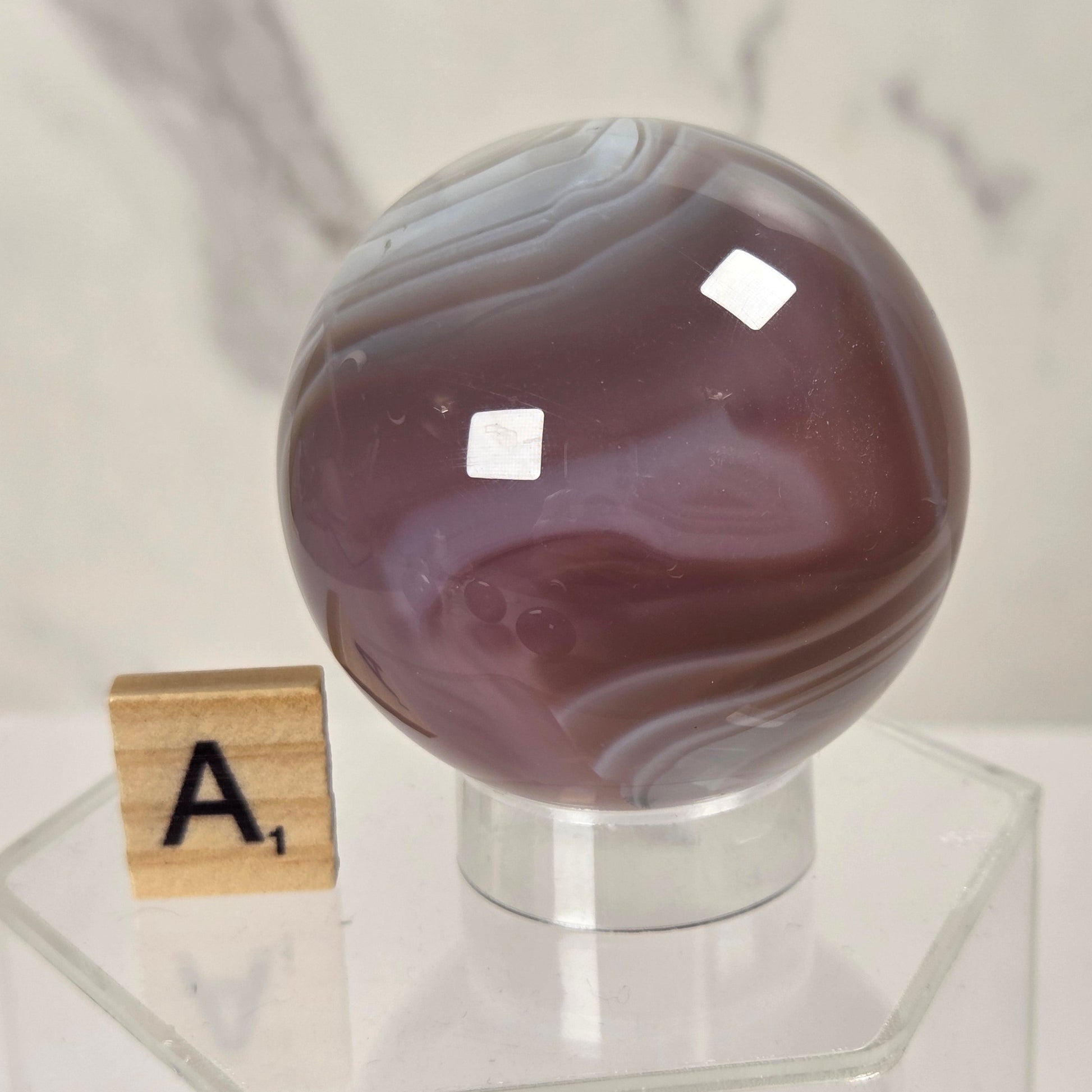 A polished round agate crystal sphere with marbled purple, gray, and white bands, displayed on a clear acrylic stand next to a wooden Scrabble tile showing the letter A.