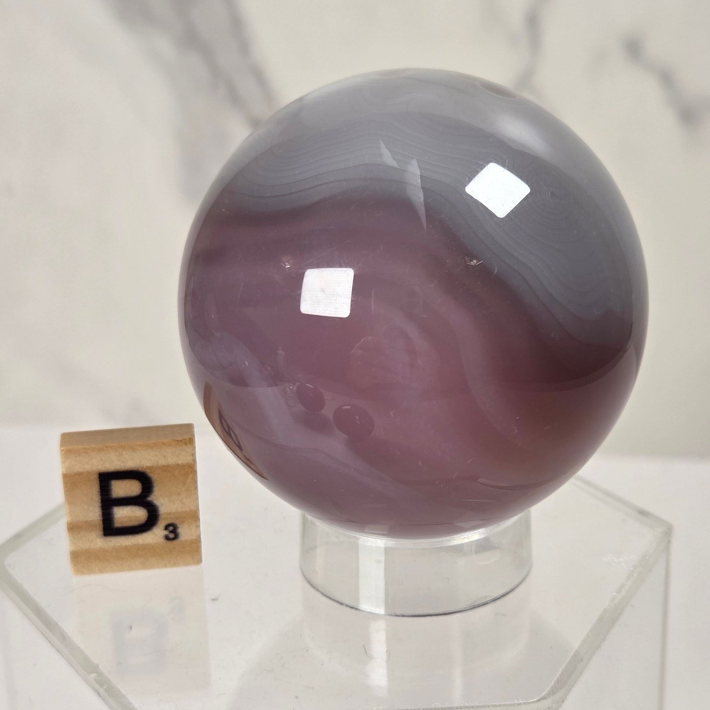 A polished purple agate crystal sphere with natural banding, displayed on a clear acrylic stand next to a wooden Scrabble tile showing the letter 'B'.
