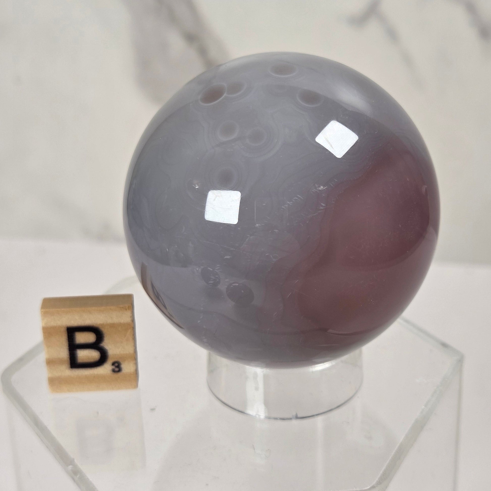 A polished oval stone display piece in smoky gray and muted mauve tones, shown on a clear acrylic stand next to a small Scrabble tile marked B³, against a light marble-pattern backdrop.