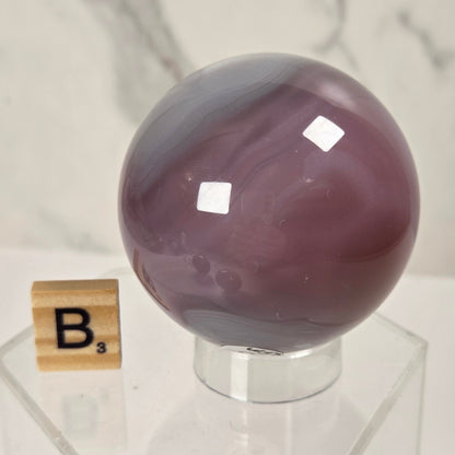 A polished purple agate crystal sphere displayed on a clear acrylic stand, with a small wooden Scrabble tile showing the letter B next to it, set against a light marble background.