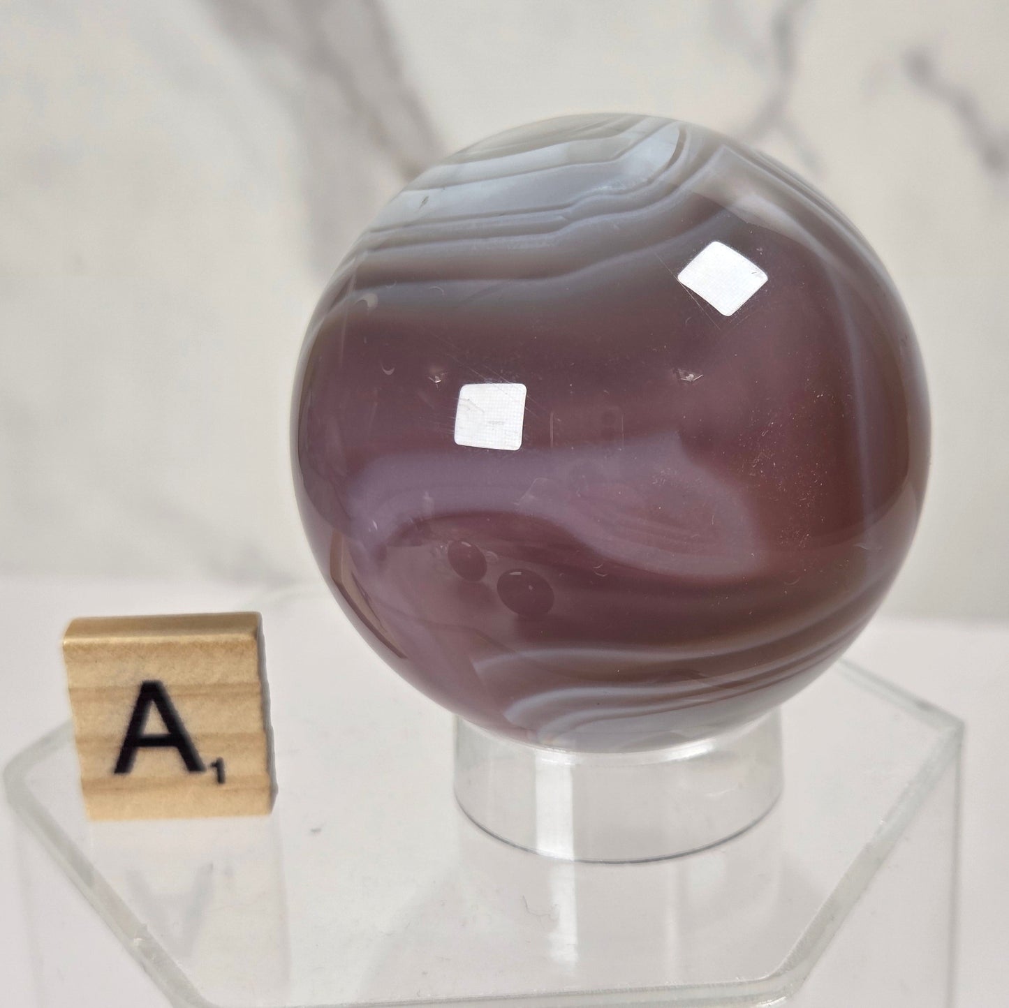 A polished round agate crystal sphere with purple and gray banding, displayed on a clear acrylic stand next to a wooden Scrabble tile on a white marble surface.