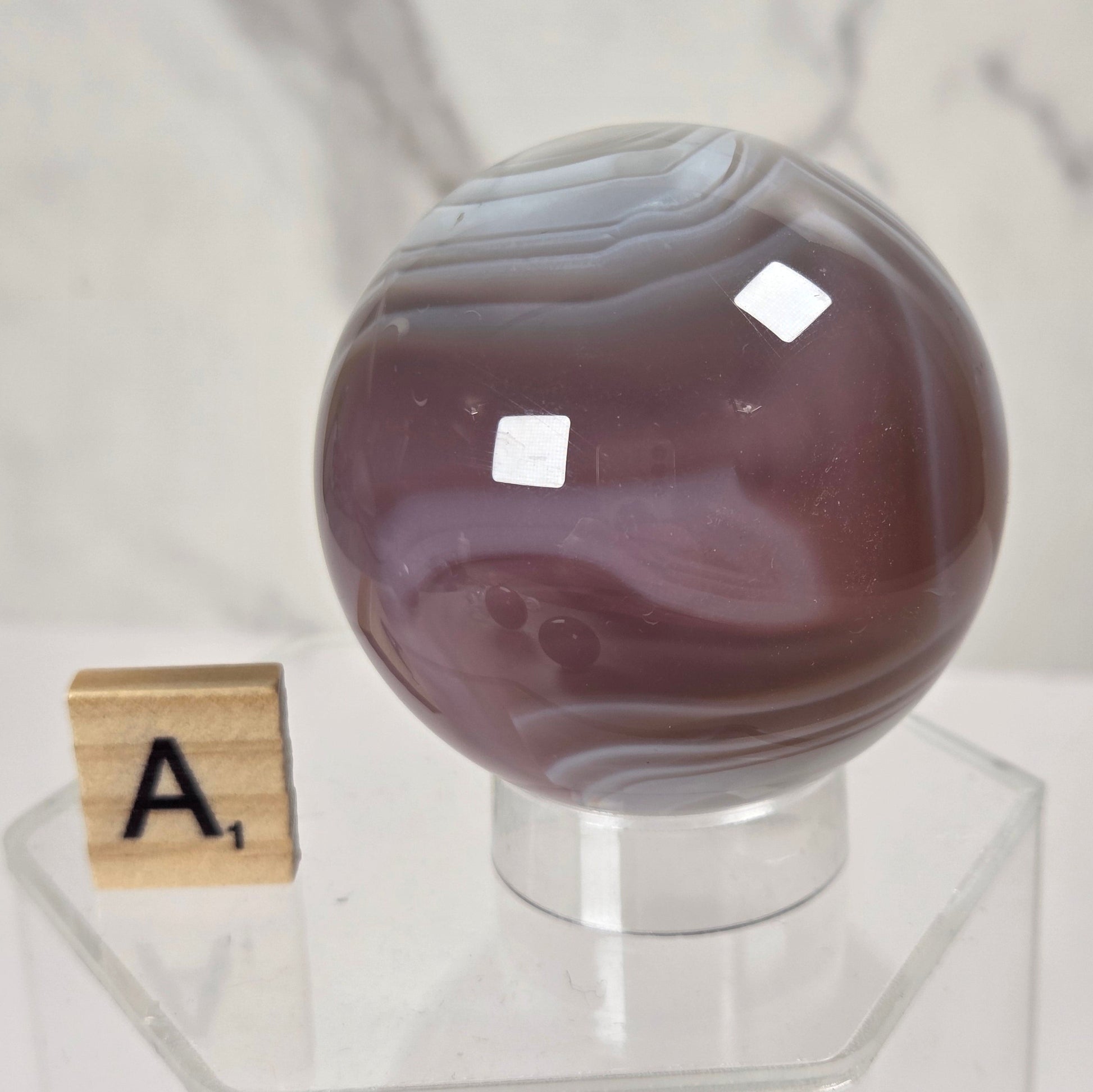A polished round agate crystal sphere with purple and gray banding, displayed on a clear acrylic stand next to a wooden Scrabble tile on a white marble surface.