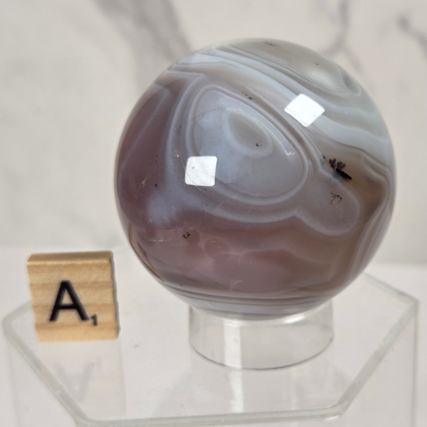 A polished agate stone sphere with swirling gray, white, and muted mauve-brown bands, displayed on a clear acrylic stand next to a small Scrabble tile showing the letter A.