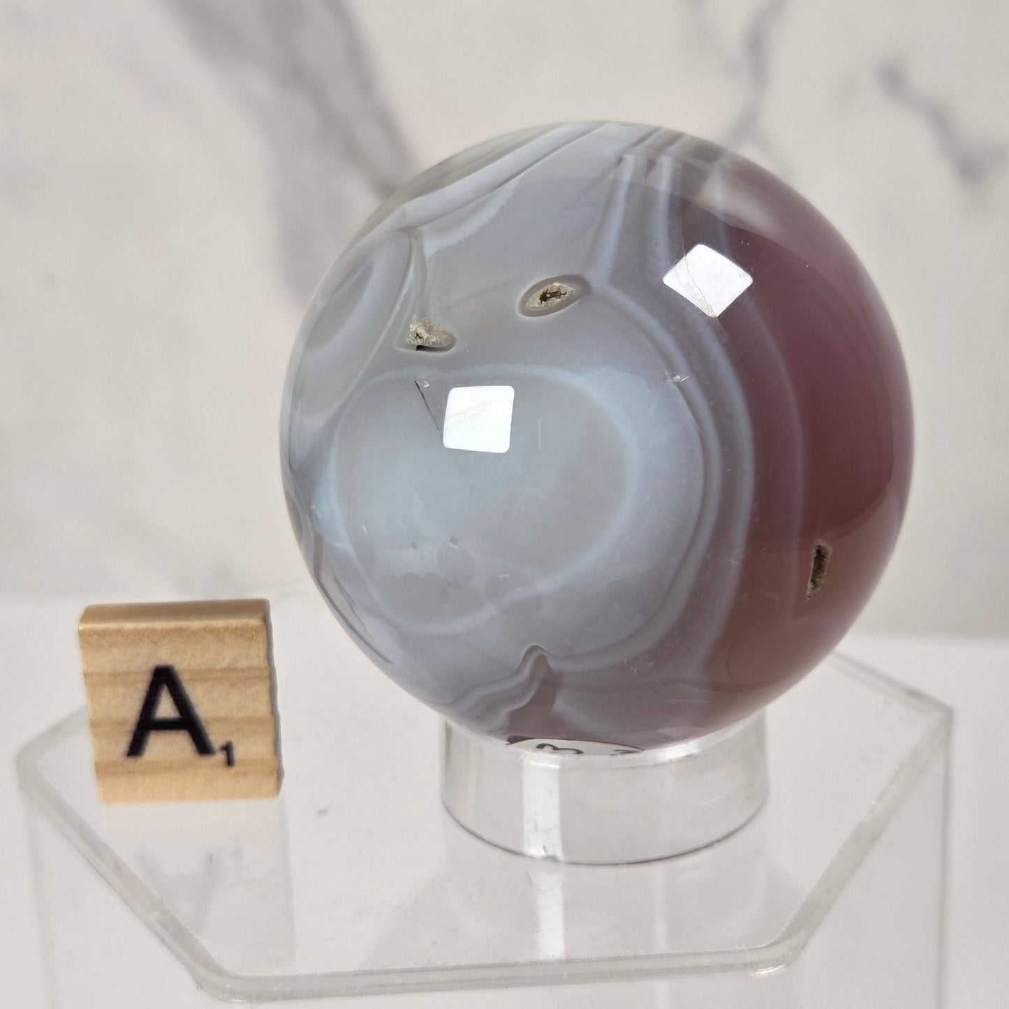 A polished agate crystal sphere with swirling gray, white, and dark reddish-brown bands, displayed on a clear acrylic stand next to a wooden Scrabble tile.