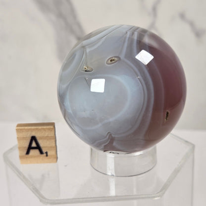 A polished agate crystal sphere with swirling gray, white, and dark reddish-brown bands, displayed on a clear acrylic stand next to a wooden Scrabble tile.