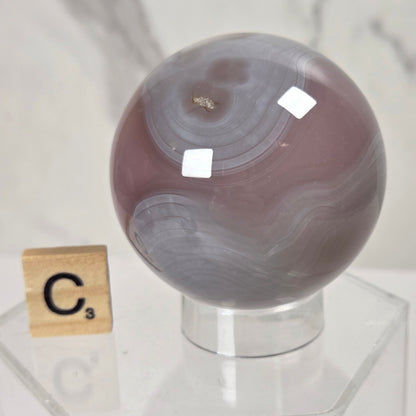 A polished agate crystal sphere with swirling gray, white, and muted mauve-brown bands, displayed on a clear acrylic stand next to a wooden Scrabble tile showing the letter C.