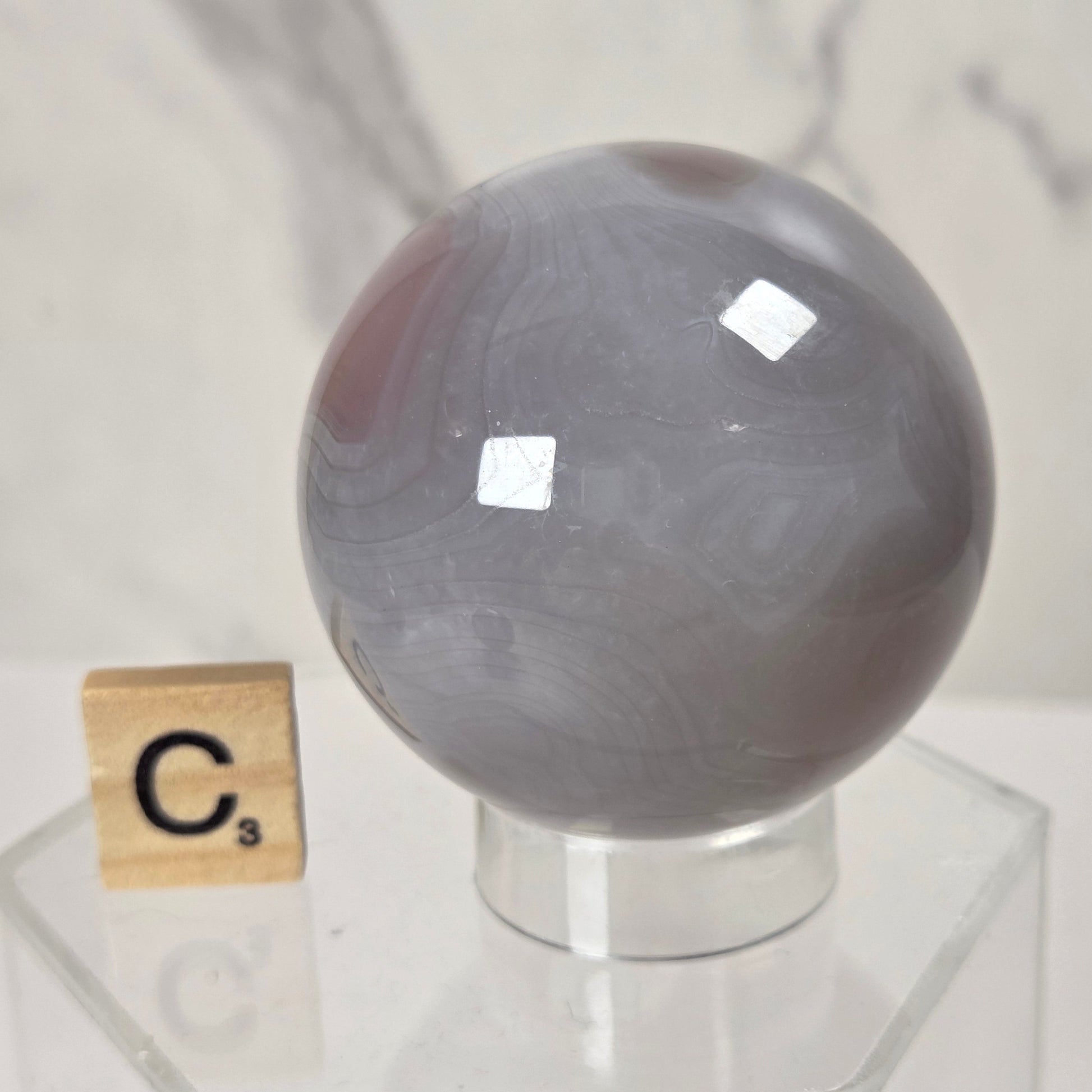 A polished gray agate crystal sphere on a clear acrylic display stand, with a small wooden Scrabble tile showing the letter 'C' placed beside it for scale, against a light marble-patterned background.