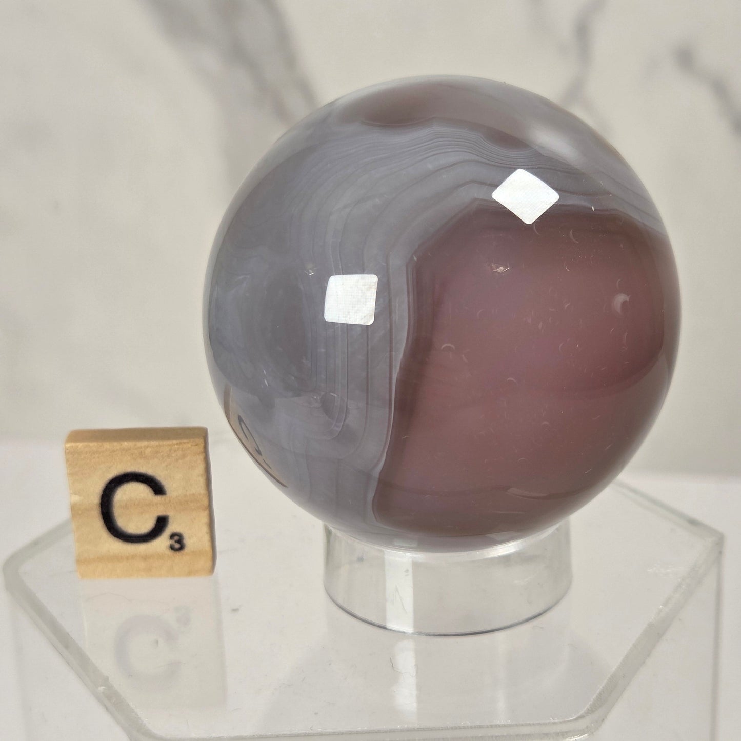A polished oval agate stone with a split gray and dusty mauve-brown coloration, displayed on a clear acrylic stand next to a Scrabble tile spelling 'C3' against a marble-patterned background.