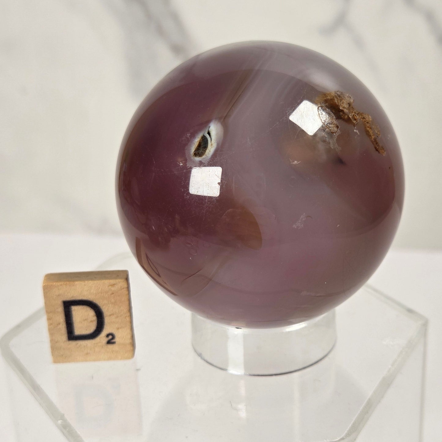 A polished purple amethyst geode sphere on a clear acrylic display stand, with a small wooden Scrabble tile showing the letter 'D' placed beside it against a light marble-patterned background.