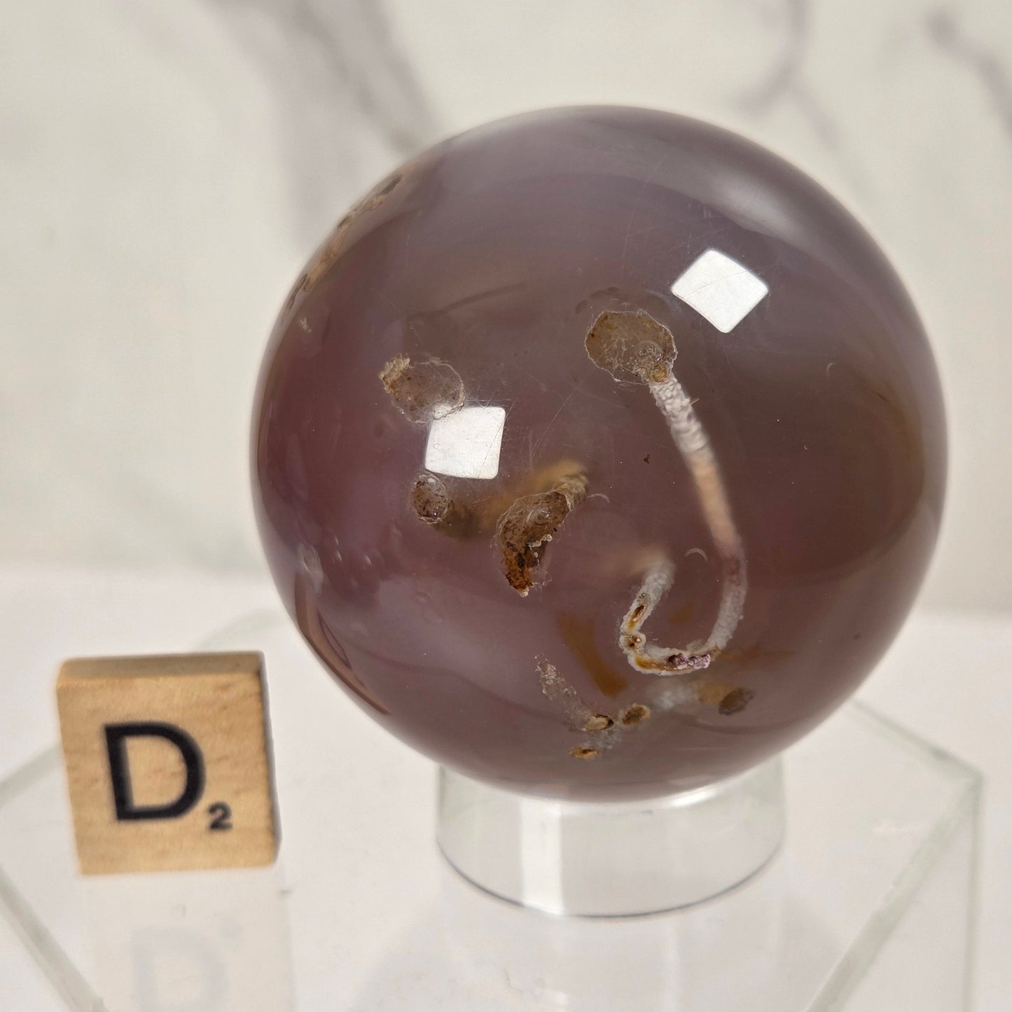 A polished round agate stone sphere with natural inclusions, displayed on a clear acrylic stand next to a small Scrabble tile showing the letter D.