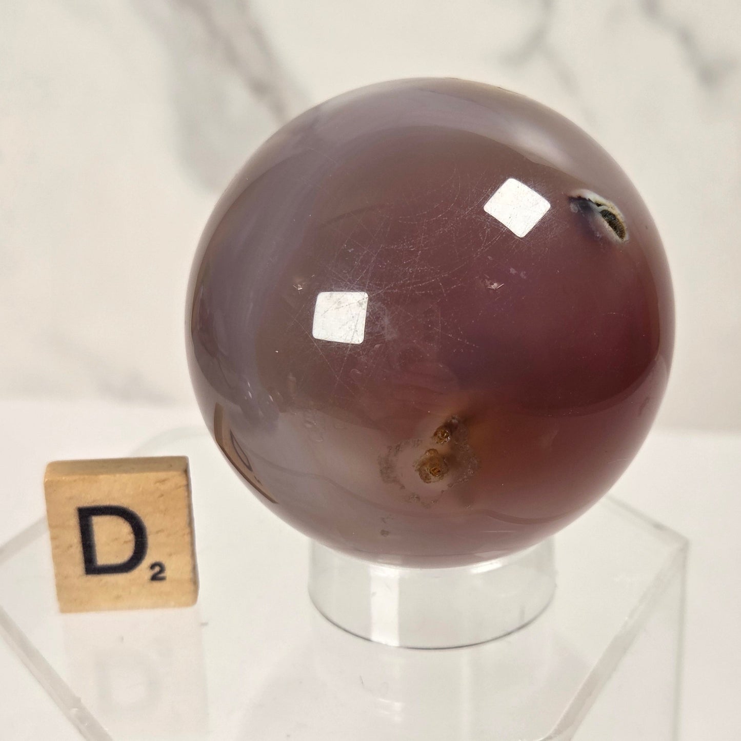 A polished oval agate crystal sphere with a deep reddish-brown hue, displayed on a clear acrylic stand next to a wooden Scrabble tile spelling 'D2' against a light marble background.