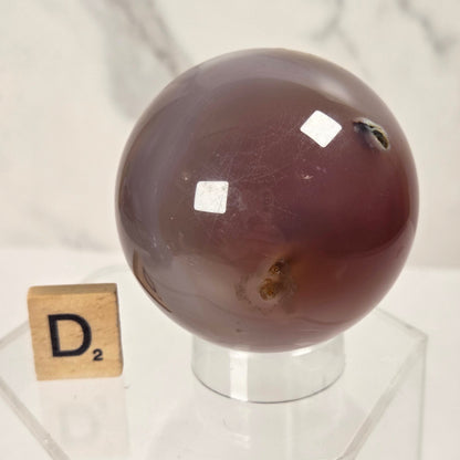 A polished oval agate crystal sphere with a deep reddish-brown hue, displayed on a clear acrylic stand next to a wooden Scrabble tile spelling 'D2' against a light marble background.