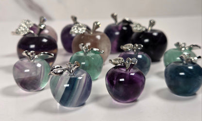 A group of polished apple-shaped jewelry pendants or charms in assorted gemstone colors, including purple, teal, blue, and smoky gray, each topped with a silver-toned leaf and small clear stones, arranged on a light surface.