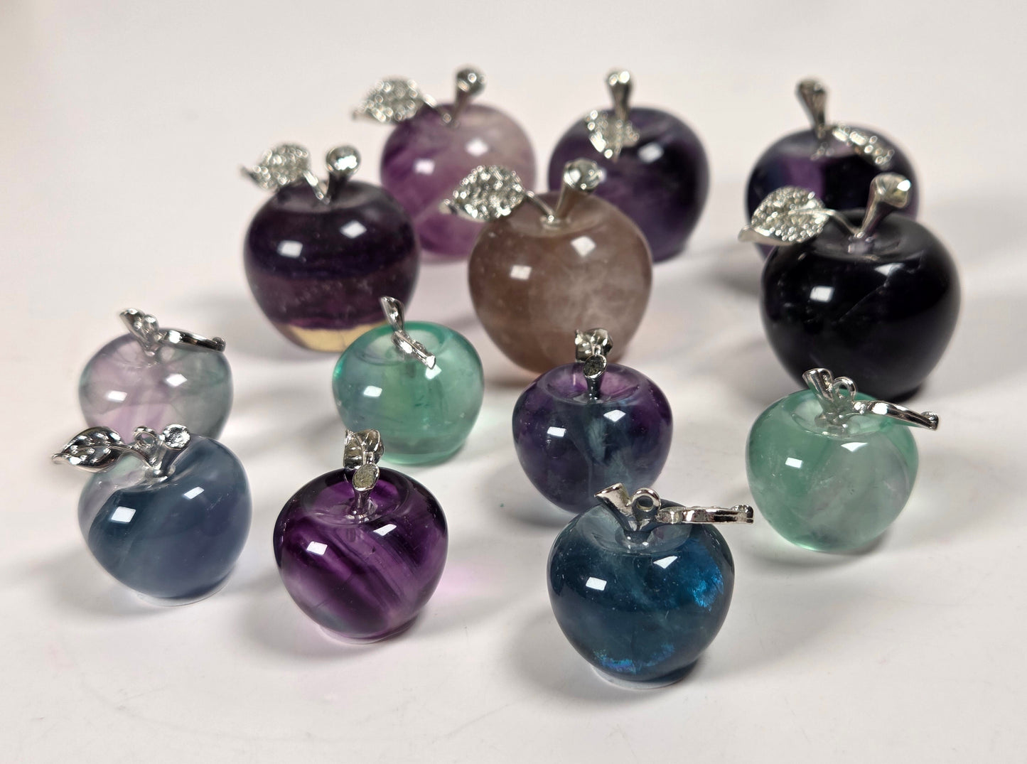 A group of small apple-shaped gemstone pendants or charms with silver-toned metal stems, displayed on a plain light background. The stones come in multiple colors including purple, blue, green, black, and pinkish-tinted varieties.
