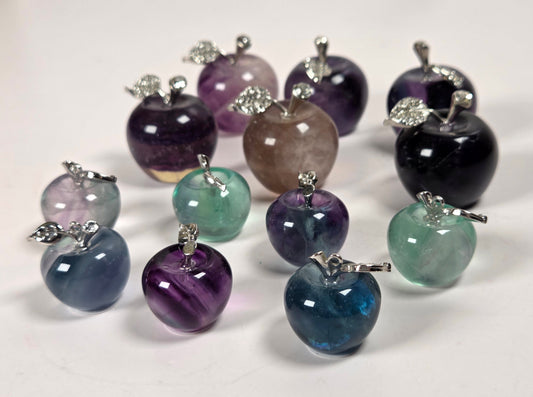 A group of small apple-shaped gemstone pendants or charms with silver-toned metal stems, displayed on a plain light background. The stones come in multiple colors including purple, blue, green, black, and pinkish-tinted varieties.
