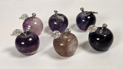 Six polished stone apple ornaments in shades of purple, black, and brown, each topped with a silver metal stem and leaf with clear rhinestones, arranged on a white marble surface.