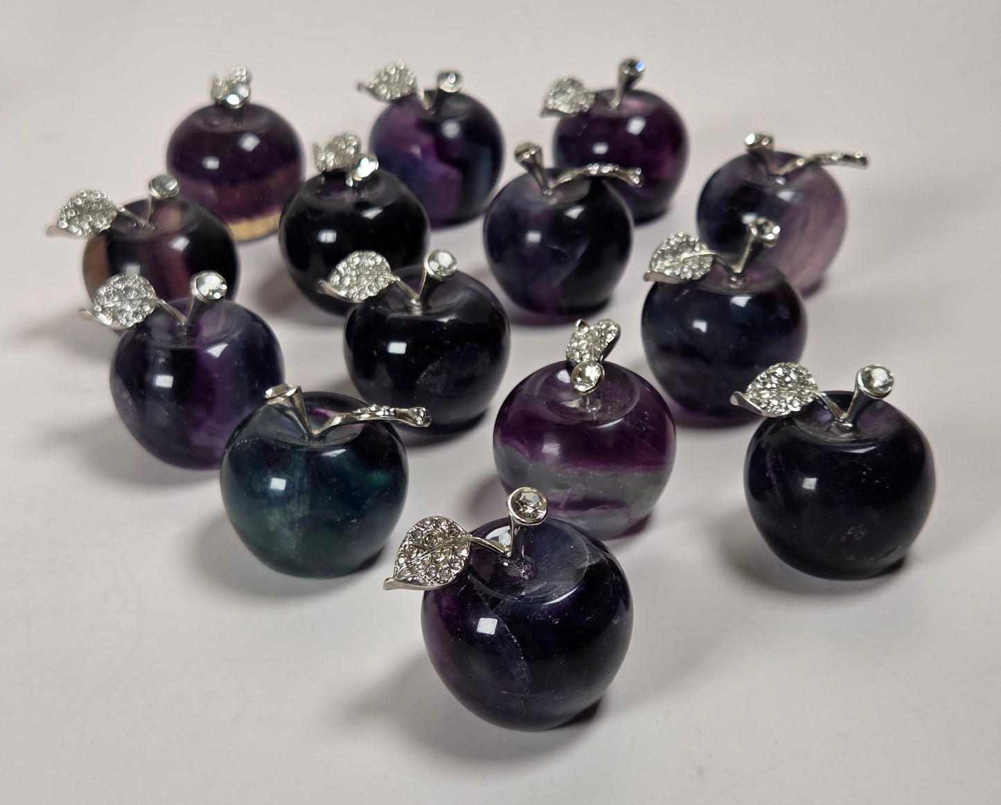 A group of small apple-shaped jewelry charms or pendants made from dark purple and black marbled stone, each topped with a silver-tone metal stem and leaf detail, arranged on a white surface.
