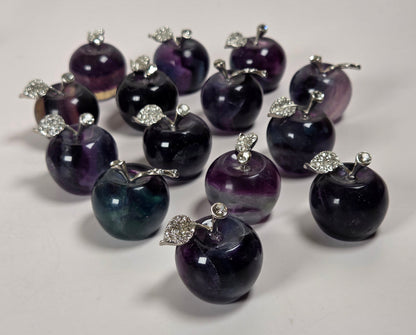A group of small apple-shaped jewelry charms or pendants made from dark purple and black marbled stone, each topped with a silver-tone metal stem and leaf detail, arranged on a white surface.