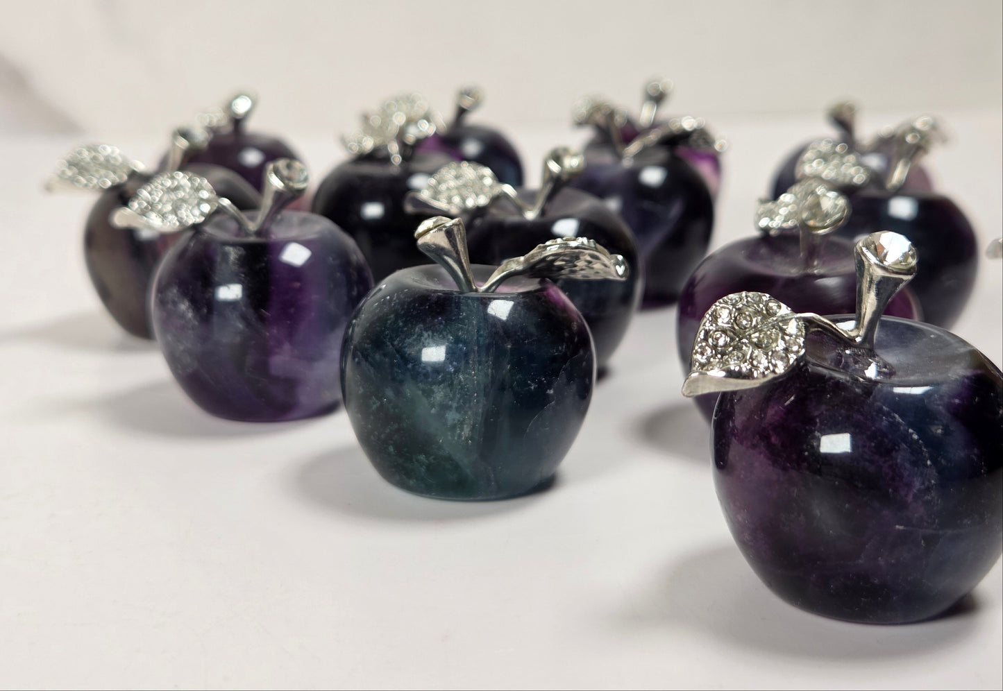 A group of small apple-shaped jewelry pieces made from polished dark purple and blue stone, each topped with a silver-toned metal stem and leaf embellished with clear rhinestones, arranged on a light neutral surface.