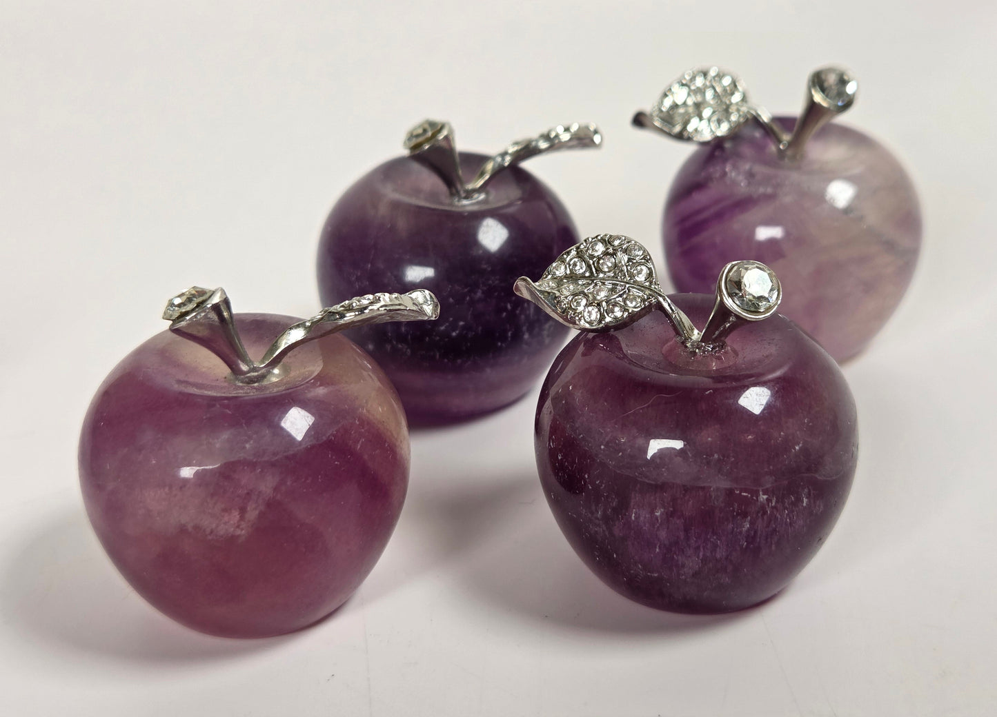 Four small apple-shaped jewelry pieces made of polished purple stone, each with a silver-toned stem and a rhinestone-encrusted leaf, arranged on a white surface.