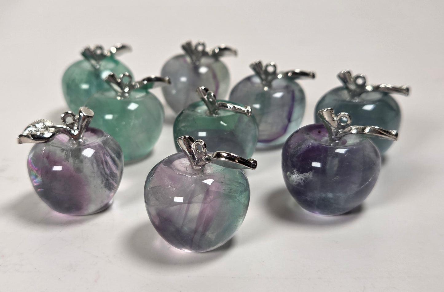 A group of small apple-shaped pendant charms made from smooth, polished gemstone showing lavender and green marbling, each topped with a silver-colored metal stem and leaf detail, arranged on a plain white surface.