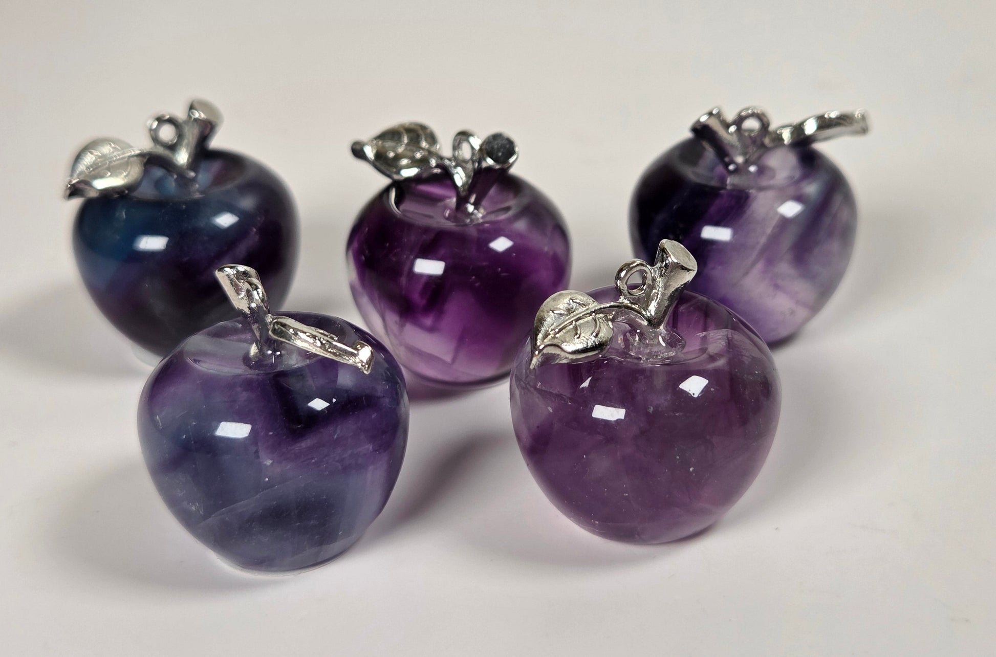 Five small apple-shaped pendants made of polished purple and blue marbled stone, each with a silver-tone metal stem detail, arranged on a plain light background.