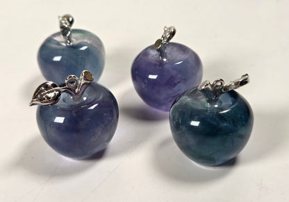 Four glossy apple-shaped gemstone pendants in shades of blue, purple, and teal, each topped with a small silver bail and leaf detail, arranged on a white surface.