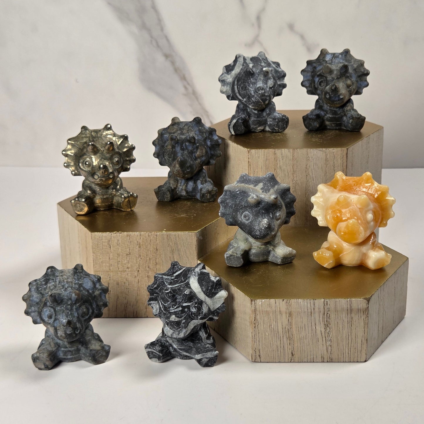 Seven small carved stone triceratops  figurines in various colors and textures, arranged on a white surface with gold hexagonal pedestals against a white marble-patterned background.
