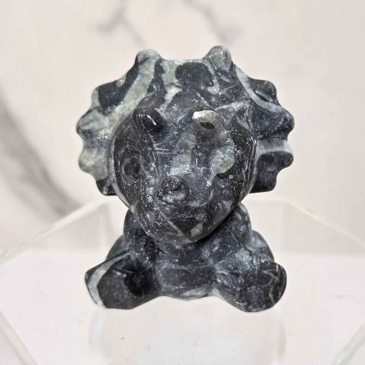 A small dark gray stone figurine with a textured, organic surface, displayed on a clear acrylic hexagonal stand against a light gray marble-patterned background.