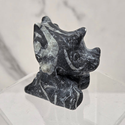 A small carved stone figurine with a dark gray and black marbled pattern, displayed on a clear acrylic hexagonal pedestal against a light gray marble-patterned background.