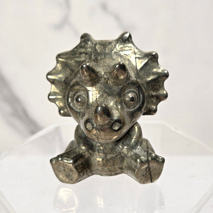 A small metallic triceratops dinosaur figurine with a textured silver finish, displayed on a clear acrylic stand against a light gray marble-patterned background.