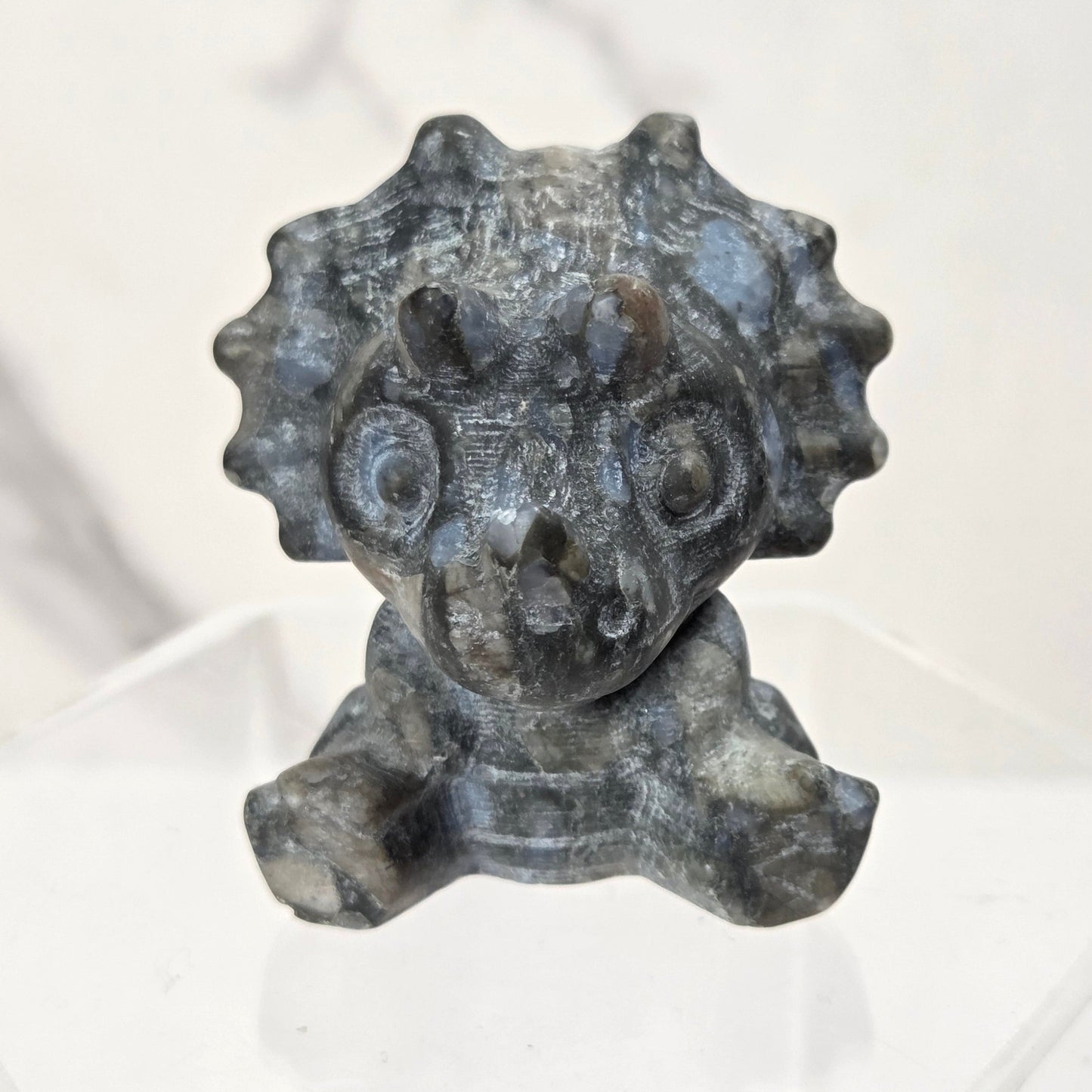 A small carved stone figurine of a cartoonish creature with a round face, large eyes, and a spiky crown, displayed on a clear acrylic stand against a light marble-patterned background.