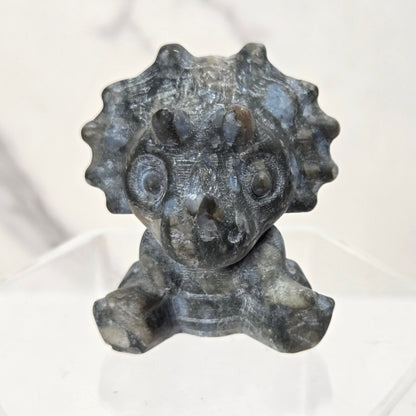 A small carved stone figurine of a cartoonish creature with a round face, large eyes, and a spiky crown, displayed on a clear acrylic stand against a light marble-patterned background.