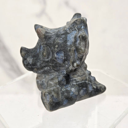A small dark gray stone figurine with blue-gray veining, displayed on a clear acrylic hexagonal pedestal against a light marble-patterned background.
