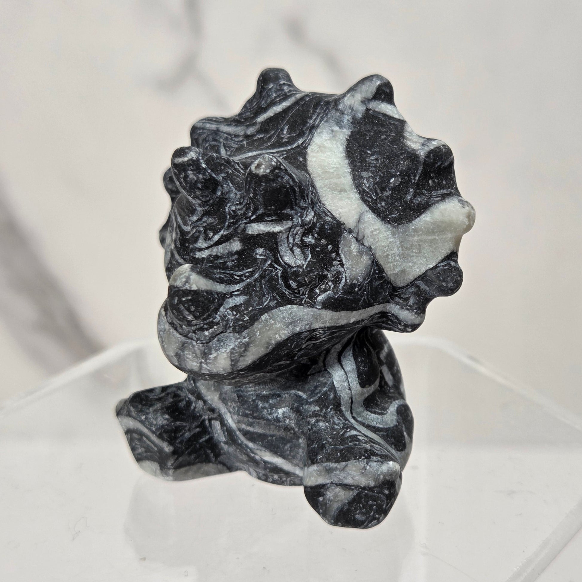 A small sculptural stone object with a marbled black and white pattern, displayed on a clear acrylic pedestal against a light marble-look background.
