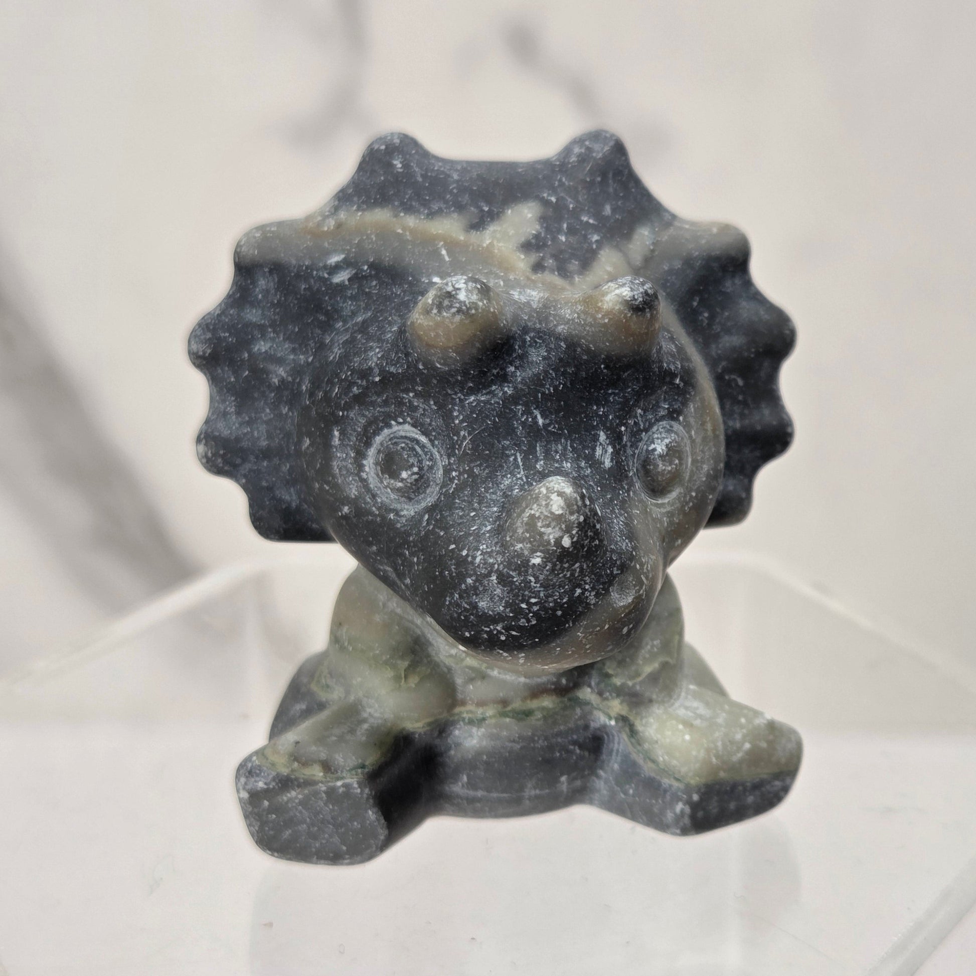 A small dark gray and black stone figurine of a seated creature with a spiky crown, displayed on a clear acrylic pedestal against a light marble-patterned background.