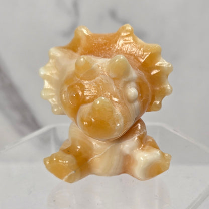 A small carved figurine of a lion or beast with a mane, made from polished amber-brown and cream stone, displayed on a clear acrylic stand against a light gray marble-patterned background.