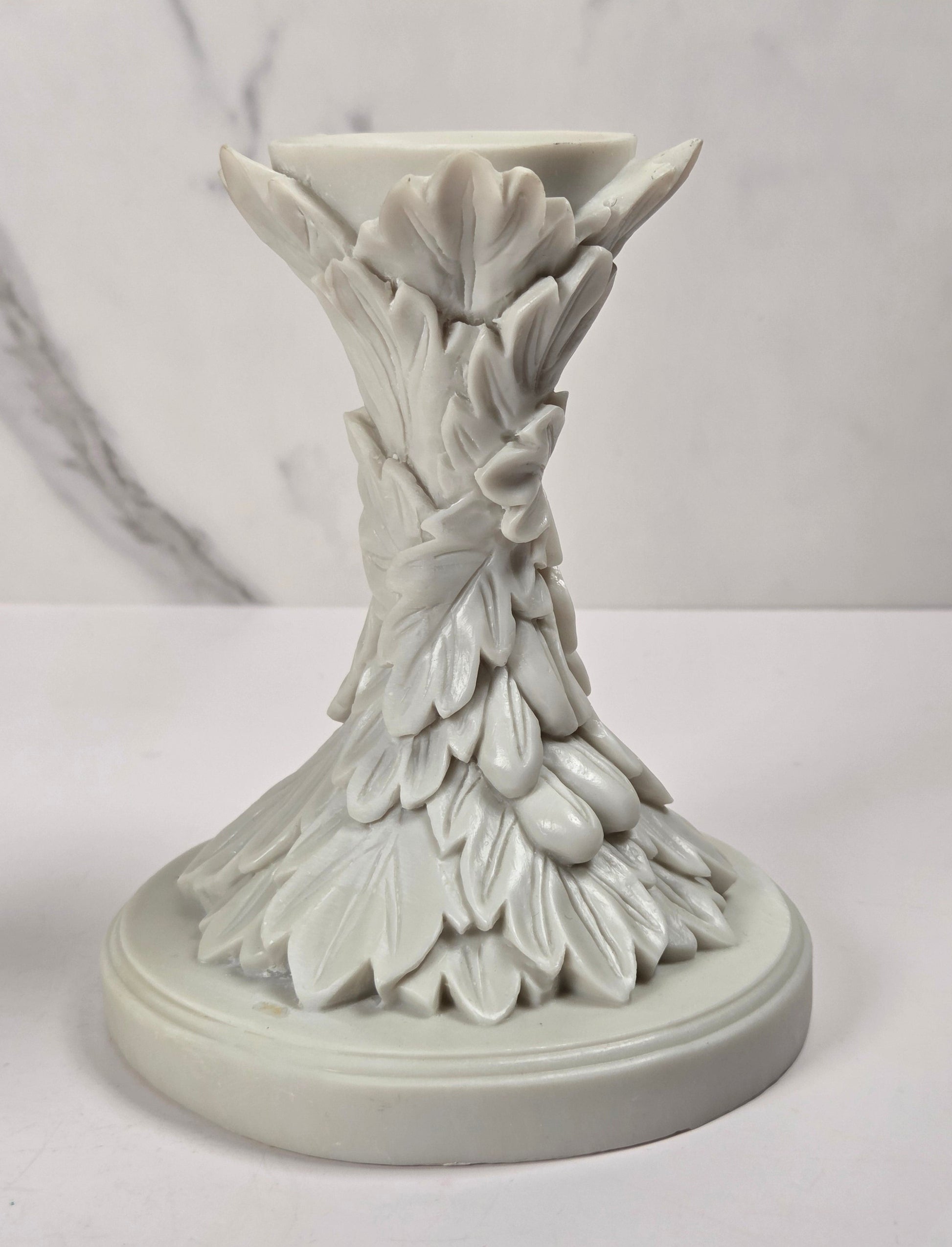 A white ceramic candle holder with an intricate, sculpted leaf and vine design on a round base, set against a light gray marble-patterned background.