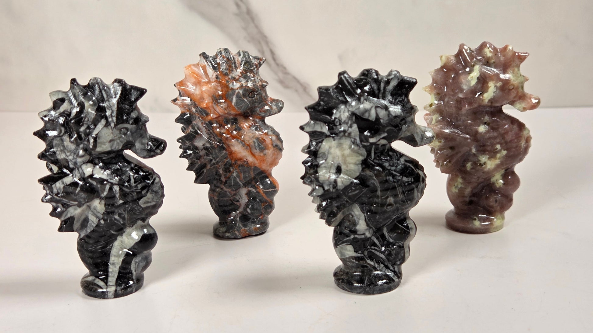 Four polished stone seahorse figurines displayed on a light surface against a marble-patterned background. The figurines are made from different stones, including black and white marbled, orange and gray marbled, and reddish-brown and cream marbled varieties.