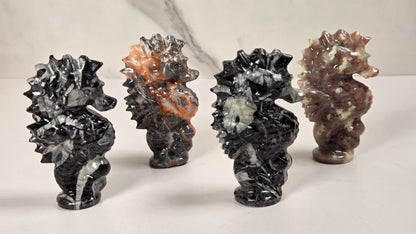 Four polished stone seahorse figurines displayed on a light surface against a marble-patterned background. The figurines are made from different stones, including black and white marbled, orange and gray marbled, and reddish-brown and cream marbled varieties.