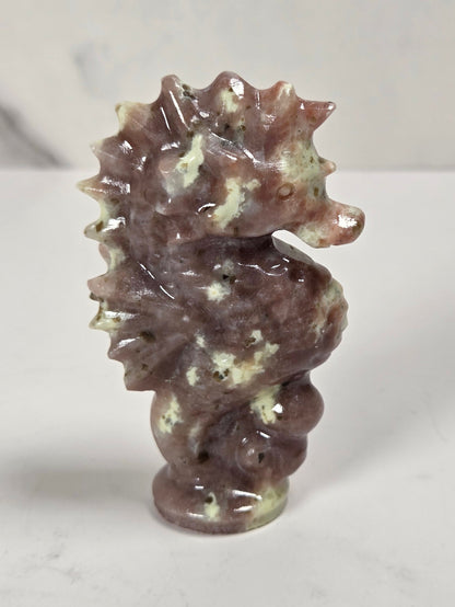 A small carved seahorse figurine made from polished brown and cream stone, standing upright on a light gray marble-like surface.