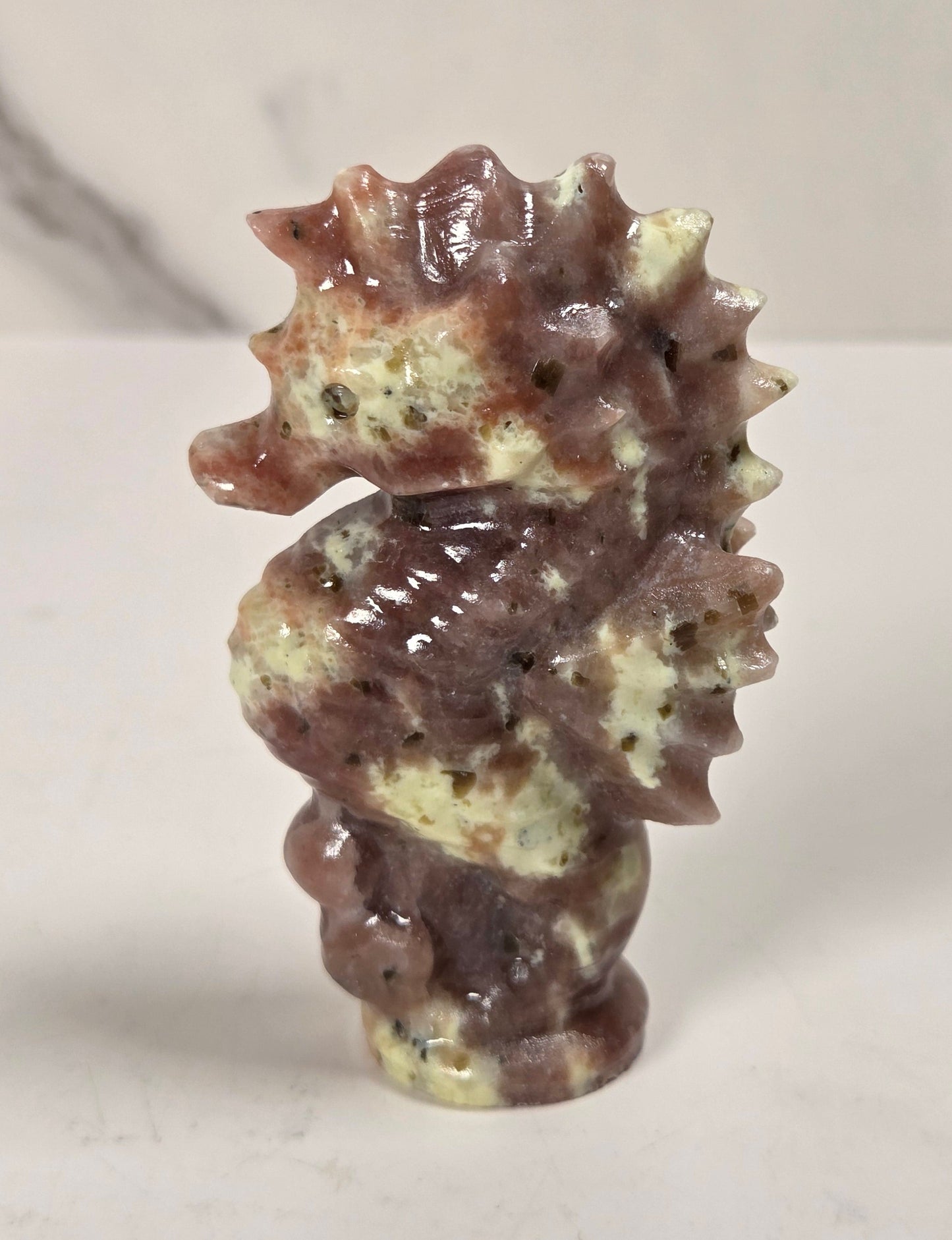 A small carved seahorse figurine made from polished stone with mottled brown, beige, and cream tones, standing on a light surface against a softly blurred marble-like background.